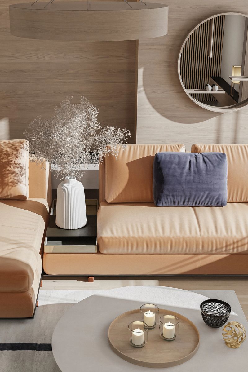 A modern luxury living room featuring a light tan leather sofa paired with a deep blue cushion for subtle contrast. A circular mirror with an integrated shelf adds architectural interest and reflects warm wood textures and curated decor. Natural light floods the space through a large window, highlighting the neutral palette and soft materials. A tall white vase with dried flowers adds a sculptural touch, enhancing the room’s calm and sophisticated ambiance. Ideal for showcasing upscale interior design and timeless residential styling.