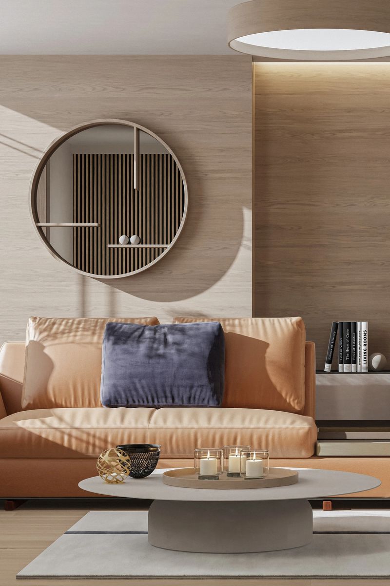 A modern luxury living room featuring a light tan leather sofa paired with a deep blue cushion for subtle contrast. A circular mirror with an integrated shelf adds architectural interest and reflects warm wood textures and curated decor. Natural light floods the space through a large window, highlighting the neutral palette and soft materials. A tall white vase with dried flowers adds a sculptural touch, enhancing the room’s calm and sophisticated ambiance. Ideal for showcasing upscale interior design and timeless residential styling.