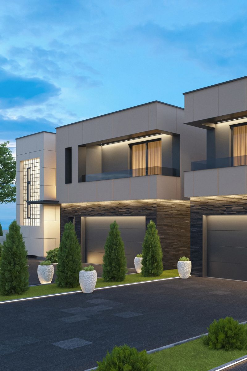 A contemporary two-story villa featuring a striking blend of dark gray stone and smooth light paneling, illuminated by warm LED accent lighting that enhances its geometric façade and clean lines. The house includes a wide double garage, a minimalist balcony, and a decorative wall with integrated lighting, all set against a serene dusk sky. The landscaped driveway is bordered by sculptural white planters and manicured evergreen shrubs, adding to the upscale ambiance. This image is ideal for showcasing premium real estate listings, architectural portfolios, or editorial content focused on modern home design, luxury living, and exterior lighting concepts.