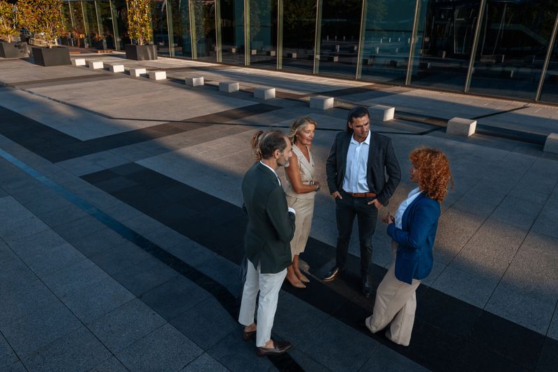A group of four adults in smart casual attire chat in an open urban space outside a glass-front building in Madrid. Collaboration, networking and professional conversation under daylight in a calm, confident atmosphere.