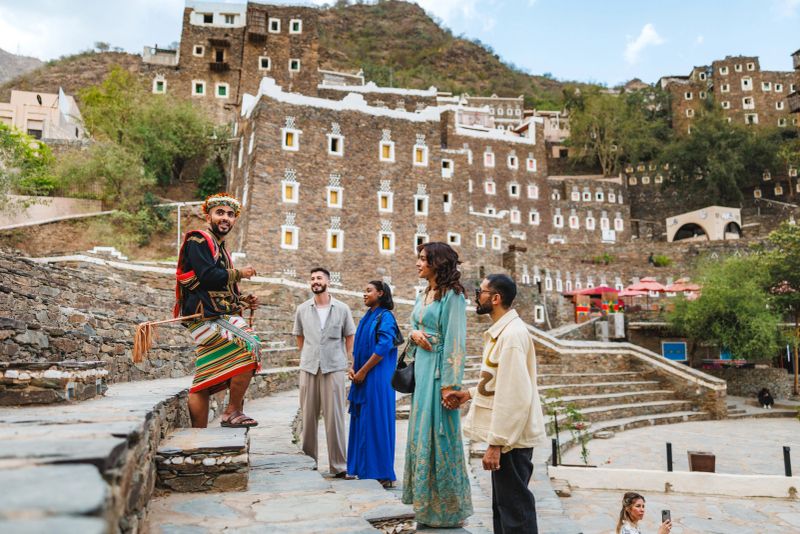 Saudi tour guide in traditional Asir dress engaging with a diverse group of tourists at Rijal Almaa amphitheater. The guide enthusiastically sharing historical stories while the group listens attentively. Traditional stone buildings and terraces provide a scenic backdrop.