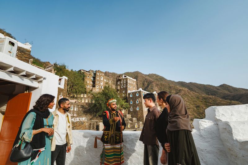Group standing with a Saudi guide in traditional Asiri dress, attentively listening. They are outdoors at the historic Rijal Almaa Mountain Village, with traditional stone buildings in the background under a clear blue sky.