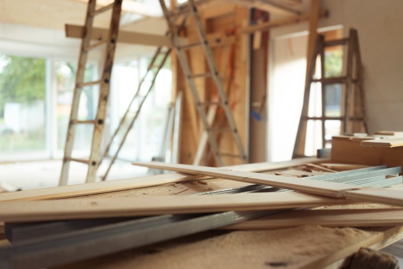 Interior of a wooden house under construction showing wooden planks, ladders, and construction materials scattered in a spacious room with large windows providing natural light, highlighting the building process