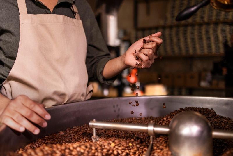 A worker gently pours and sprinkles roasted coffee beans from a large roasting tub, capturing the aroma and motion of batch roasting in a bustling cafe setting
