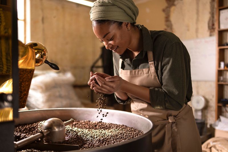 A focused woman in an apron and headscarf roasts coffee beans, pouring them into a grinder in a rustic, sunlit workspace, capturing the craft and aroma of coffee production
