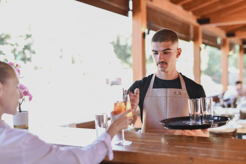A female bartender in a white shirt and waiter are preparing and hands drinking glasses for a customer at a wooden bar. Refrigerated beverages line the back wall, capturing a casual hospitality moment.