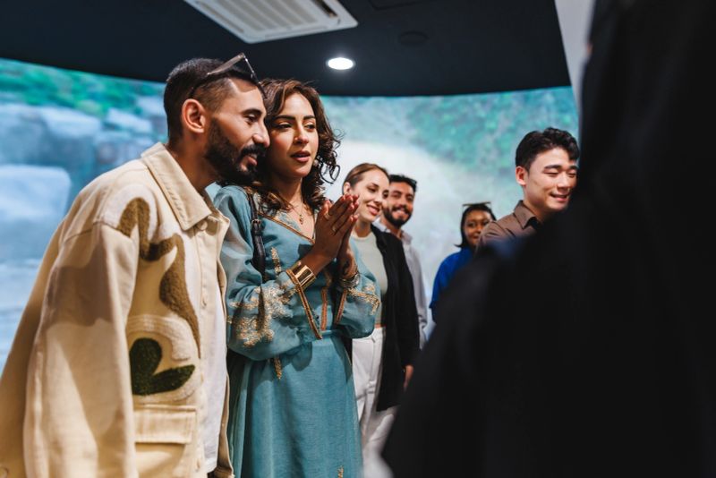 Group of diverse tourists gathered in a museum setting at Rijal Almaa. They are smiling and engaged as they listen to a guide. The environment is modern with a large digital display in the background.