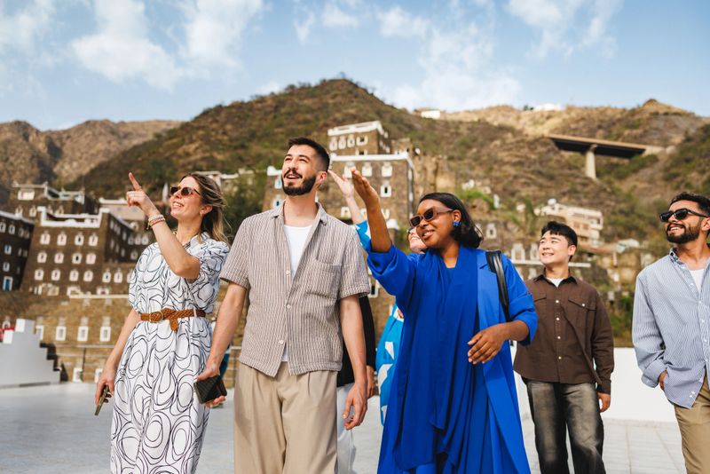 Group of friends walking through Rijal Almaa Heritage Village in the Asir Mountains. They are smiling and pointing at the historic architecture while enjoying the picturesque landscape under a bright blue sky.
