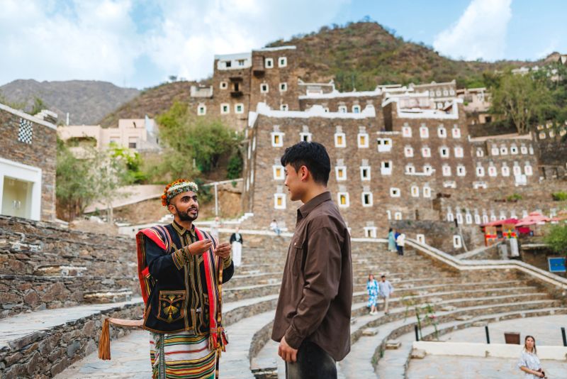 Local Saudi guide in traditional attire engaging with an Asian visitor in Rijal Almaa. They are standing outdoors with historical architecture in the background. The guide is gesturing animatedly while explaining the cultural significance of the site.
