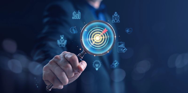 A businessman in suit holding a magnifying glass focusing on a glowing target surrounded by business icons, representing business goal, strategy, and success concept in modern digital world.