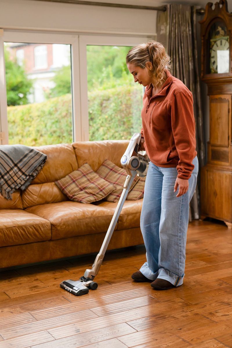 A woman is using a vacuum cleaner to tidy up a hardwood floor in a cozy living room filled with natural light, enjoying the cleaning process.
