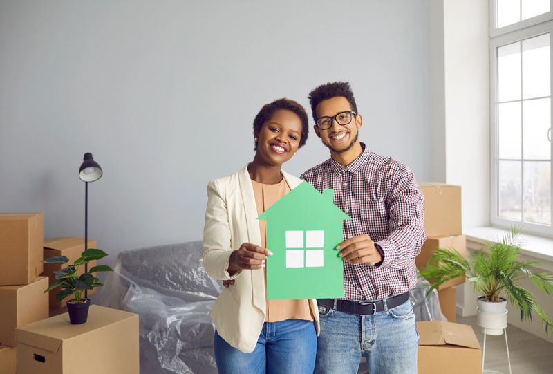 Happy black married couple in their new home. Portrait of young family standing in the living room interior, holding symbolic paper house, looking at camera and smiling. Buying real estate concept