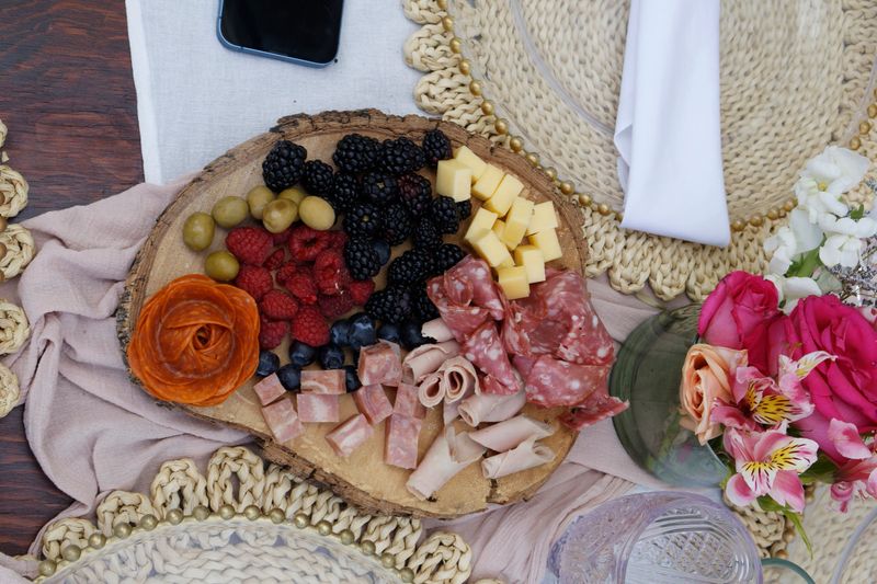 Charcuterie board features an array of cheeses, sliced meats, olives, blackberries, raspberries, and blueberries on a wooden platter. It rests on a tea party-themed table with woven placemats and a white napkin. A black smartphone is placed nearby. Fresh flowers in a vase, including pink roses, add decorative touches. The board is surrounded by light-colored fabric and a glass cup, creating an elegant and inviting setting.