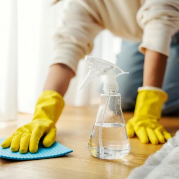 Person cleaning a wooden floor with gloves and spray bottle.