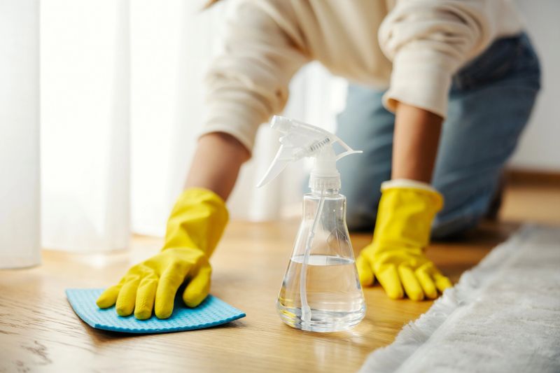 Woman wearing yellow rubber gloves cleaning a wooden floor using a spray bottle and blue microfiber cloth at home