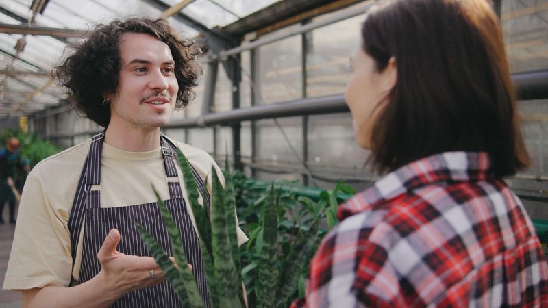 A plant shop worker talks to a customer in a greenhouse filled with various plants, creating a welcoming atmosphere for gardening enthusiasts.