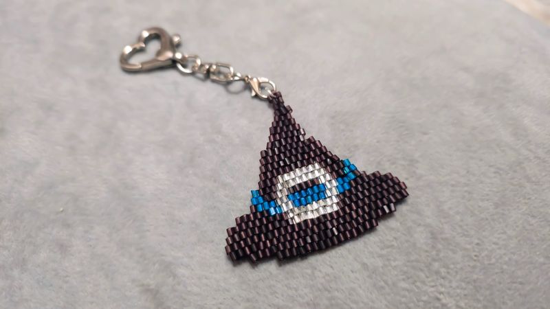 A cartoon character-shaped keychain, handcrafted from multicolored seed beads and glass beads on a gray background. Product demonstration.