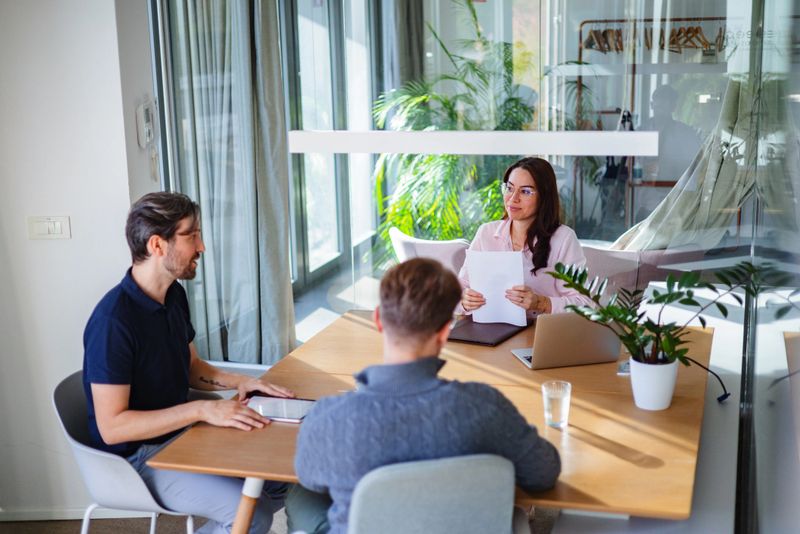 A group of business people in smart casual attire engaged in a collaborative meeting. The diverse team is focused on brainstorming ideas while seated around a desk with laptops and documents. Sunlight streams through large windows, creating a warm atmosphere.