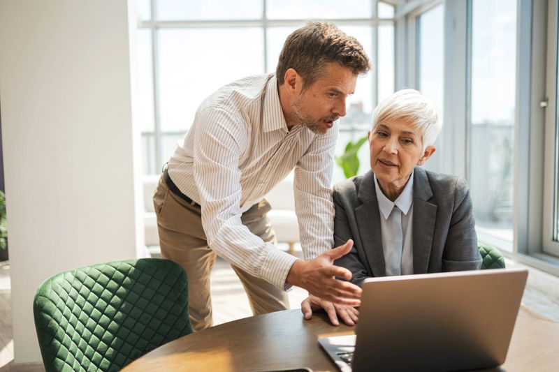 An experienced professional and a colleague collaborate at a desk with a laptop, emphasizing teamwork, communication, and professionalism within a bright and modern workplace environment.