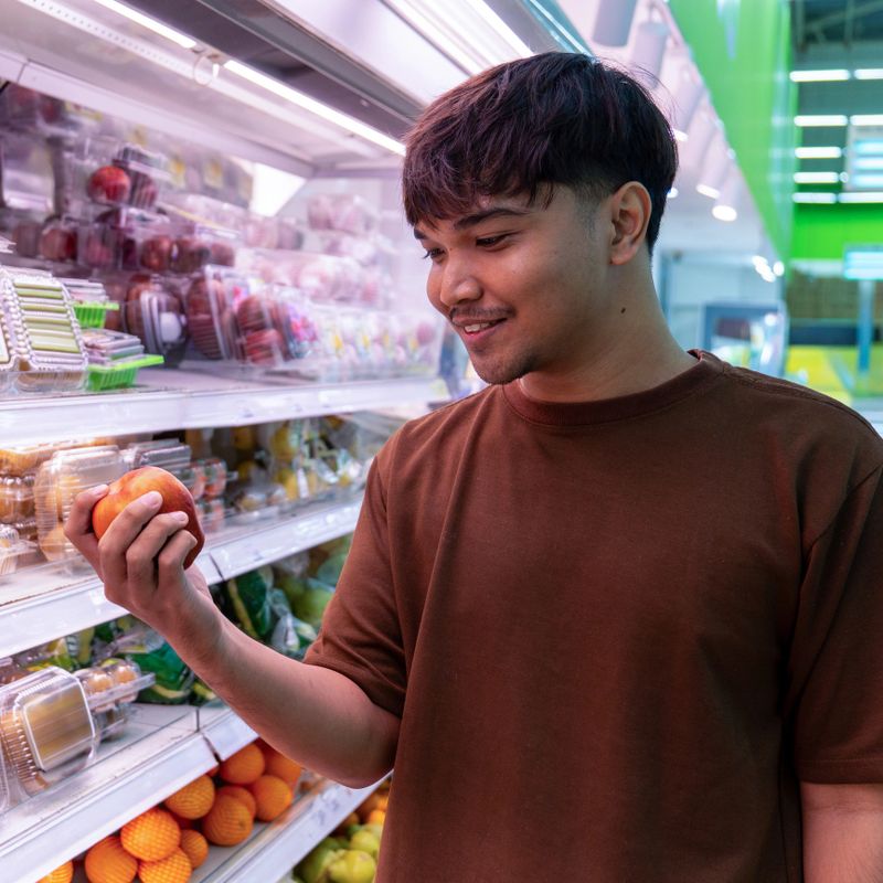 Young Asian man holding an apple while shopping for fruits in a supermarket. Healthy eating, fruit selection, and fresh produce concept.