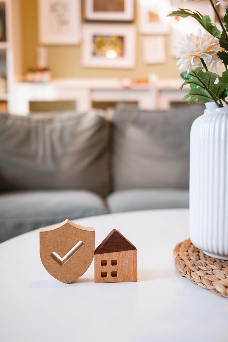 Wooden house model and shield with check mark on table in cozy living room, symbolizing home insurance, property protection, safety, and real estate security concept.