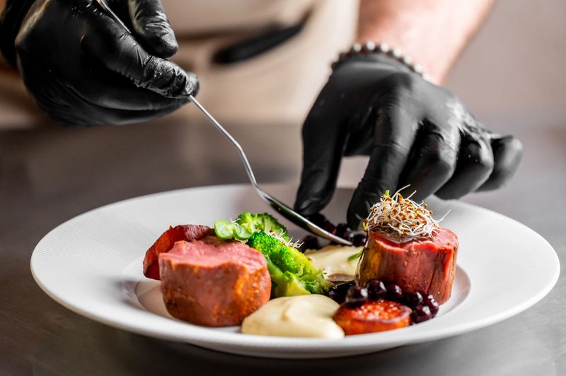A chef with black gloves carefully plates a gourmet dish of meat, broccoli, and purees on a white plate, highlighting fine dining and culinary artistry