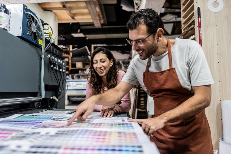 Professional colleagues collaborate in a modern digital printing office, carefully reviewing printed materials and color samples spread across a table, creating an authentic moment of teamwork in the printing and design industry.