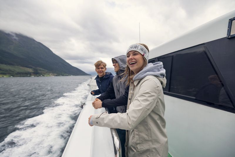 On the open deck of a fjord ferry slicing through cold northern waters, three teenage travelers stand in awe, their jackets rippling in the wind. Behind them, the boat’s wake stretches sharply across the surface. Around them, steep green mountains rise into low-hanging clouds, their peaks veiled in mist. The overcast sky casts a cool, silvery light over the scene. The group is looking at the landscape with both joy and reverence. Shot with Canon R5