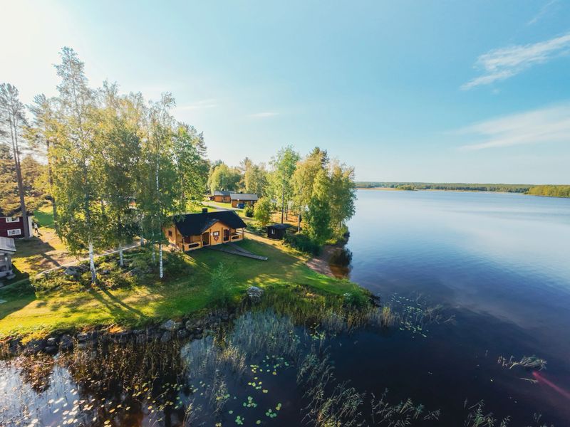 Scenic aerial view of a traditional wooden log house located on the shore of a calm Finnish lake, surrounded by dense green forest. Peaceful Nordic landscape with reflections on the water, summer cabin lifestyle, and pristine wilderness in Finland. Ideal for concepts of nature, travel, relaxation, and Scandinavian countryside living.