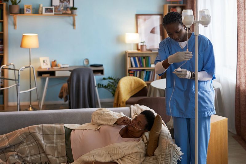 Senior Black woman lying on sofa under blanket, receiving intravenous therapy from young adult Black female nurse, wearing medical gloves, adjusting IV drip in home setting
