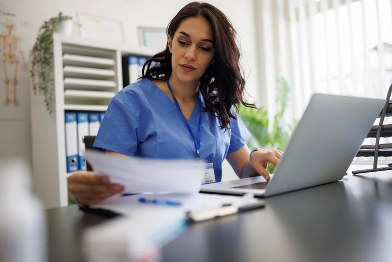 A focused female doctor or nurse wearing blue medical scrubs sits at a desk, holding paperwork while typing on a laptop computer. The healthcare professional works in a bright medical office, managing patient records and reviewing medical documentation. The image conveys professionalism, healthcare administration, and modern telemedicine concepts.