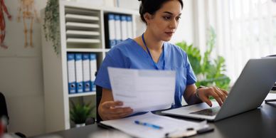 A healthcare professional reviews documents while working on a laptop in an office.