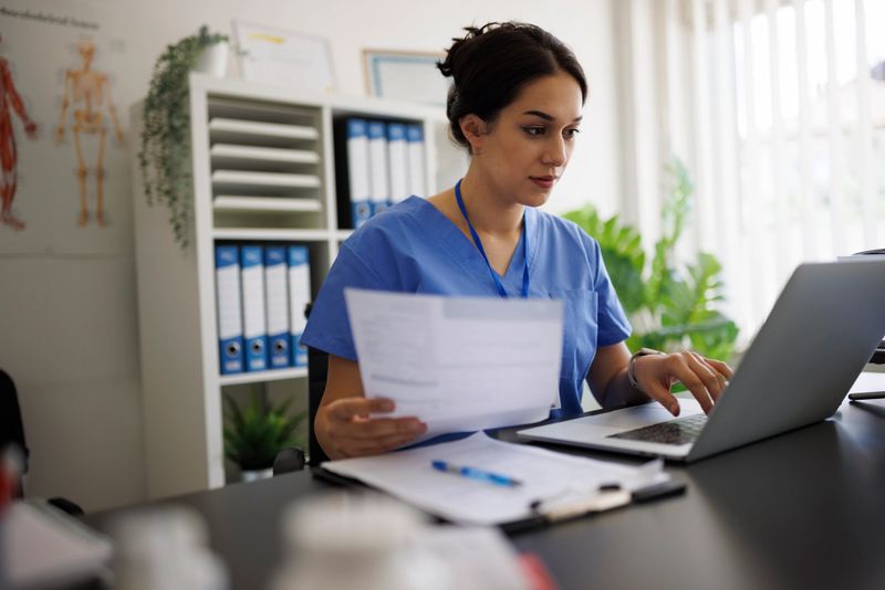Portrait of a professional female healthcare worker, either a nurse or doctor, wearing blue medical scrubs and a badge, sitting at her desk in a modern medical office. She is holding a patient's report while typing on her laptop, demonstrating focus and professionalism. The setting includes office folders, green plants, and soft daylight, creating a calm and organized medical environment. Ideal for concepts of healthcare documentation, telemedicine, clinical administration, or modern medical practice.