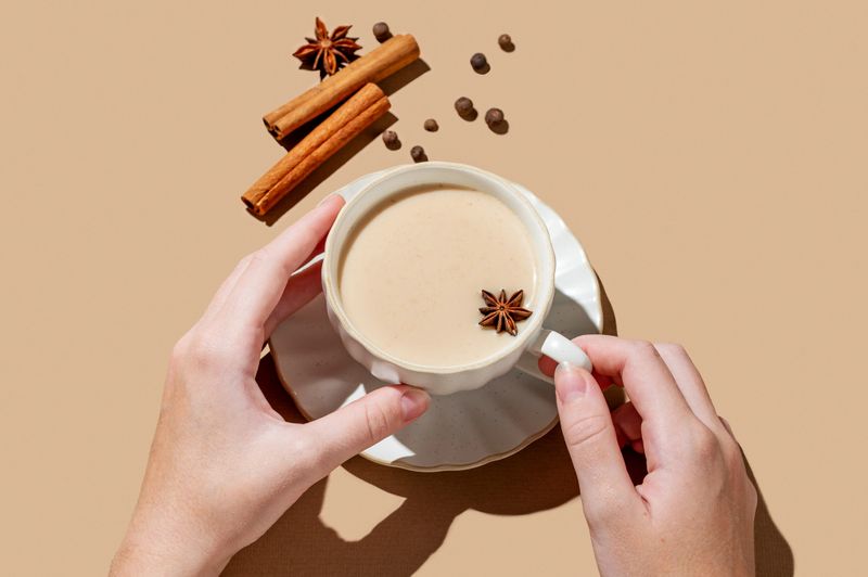 Hand holding cup of traditional Indian masala chai tea with milk and spices on a beige background with cinnamon, anise and shadows. A cozy hot drink concept for a menu or banner for an ethnic cafe or restaurant. Top view, copy space.