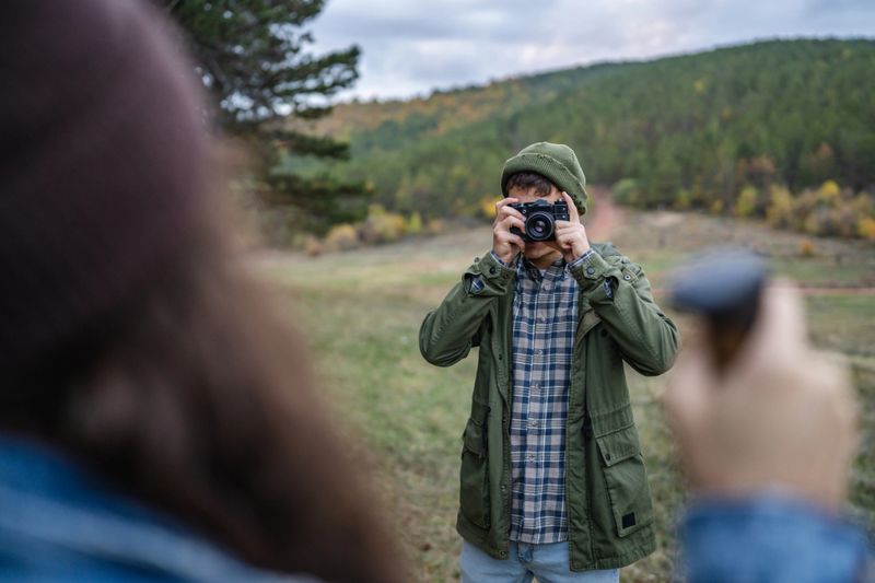 Man in casual clothing using a camera to capture a subject off-frame while standing in a natural landscape during an autumn day, with his car parked in the background