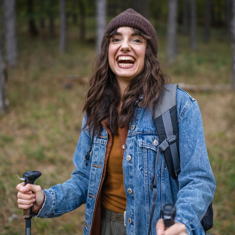 Young woman standing in a forest with backpack and hiking poles, smiling at camera while enjoying a relaxed autumn trek, feeling joyful, free and healthy outdoors