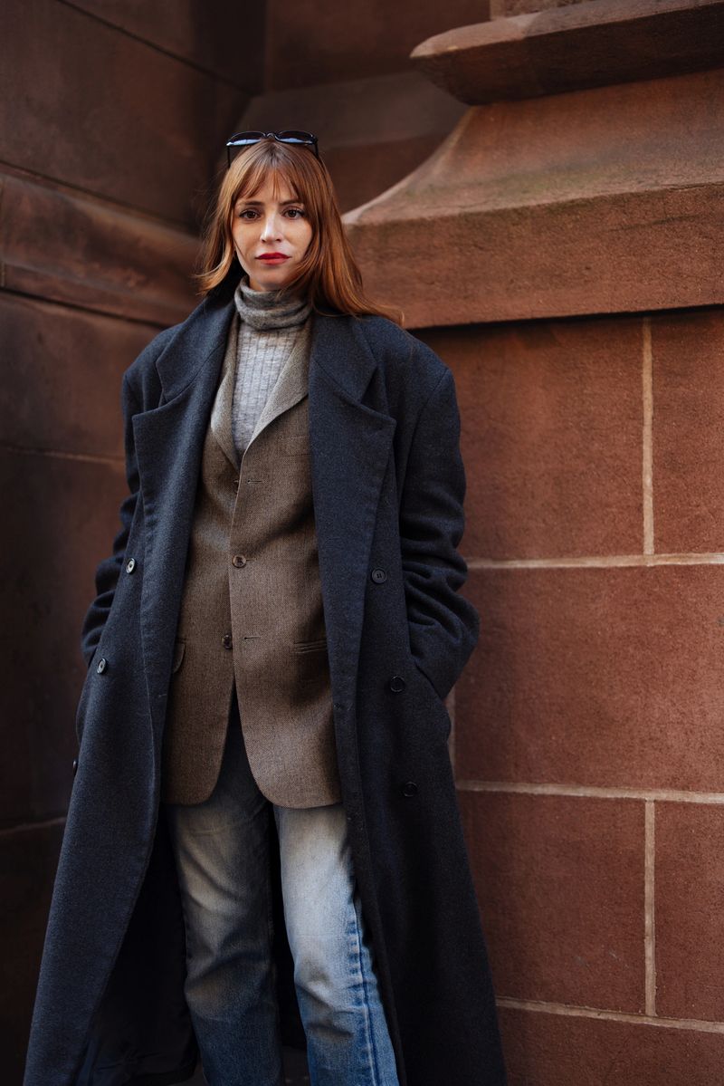 A stylish woman with ginger hair stands against a red brick city wall, wearing a long navy coat over a brown blazer and gray turtleneck. She exudes urban fashion and street confidence.