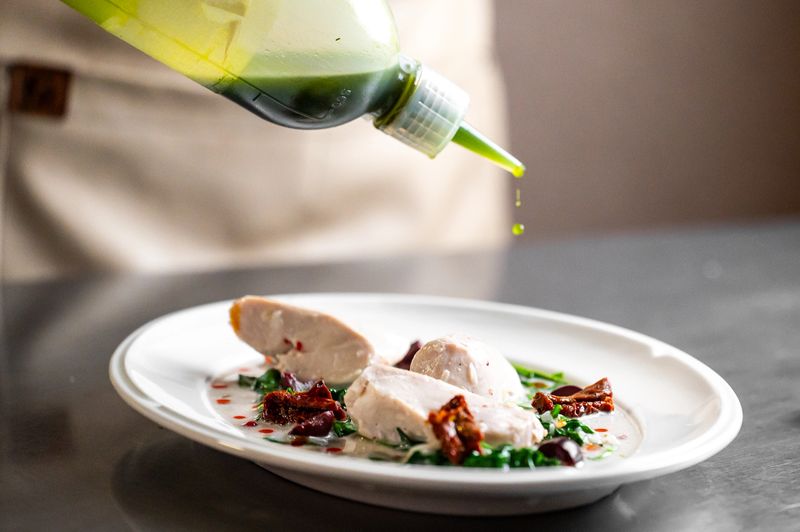 A chef carefully plates a gourmet dish of sliced chicken, a creamy sauce, and sun-dried tomatoes, drizzling a bright green oil from a squeeze bottle