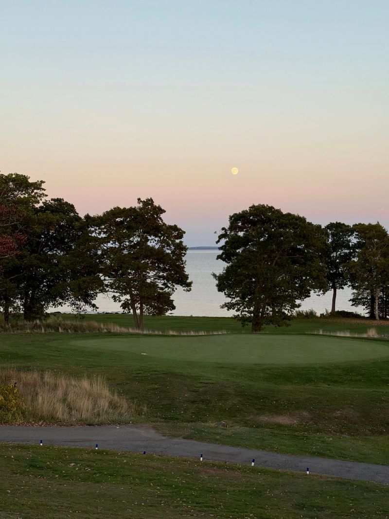 Scenic view of a golf course at sunset with a full moon rising over a calm sea, framed by trees.