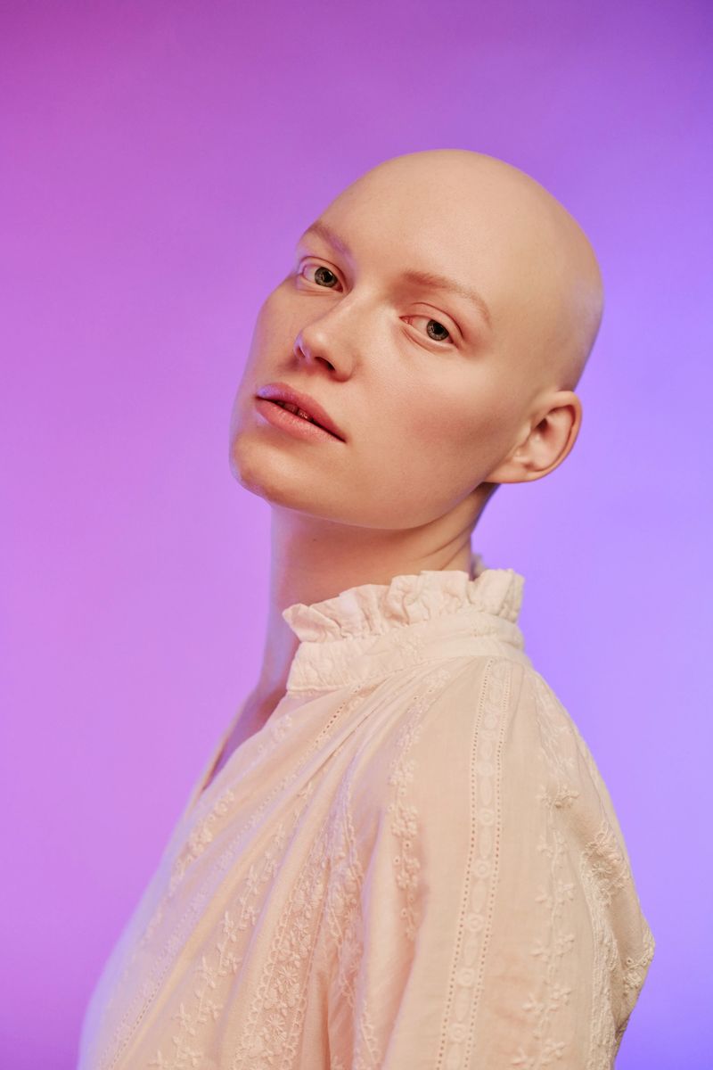 Portrait of young adult Caucasian woman with alopecia tilting head slightly and gazing into camera, showing smooth bald scalp and delicate facial features against gradient background
