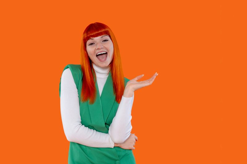 A joyful woman with bright orange hair and a green top smiles widely while posing playfully in front of a bold orange backdrop, showcasing her energetic personality.