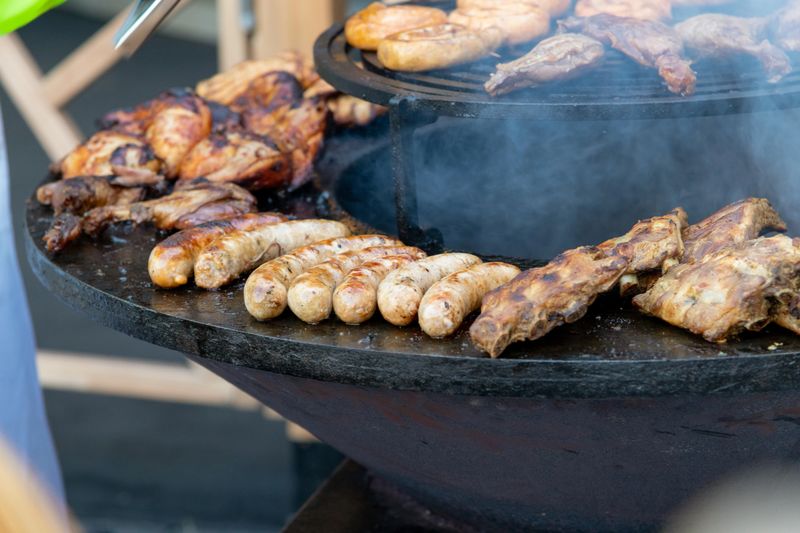Juicy sausages and marinated meats cook over an open flame at a lively barbecue gathering. Smoke rises as the grill sizzles, creating an inviting atmosphere for food lovers.
