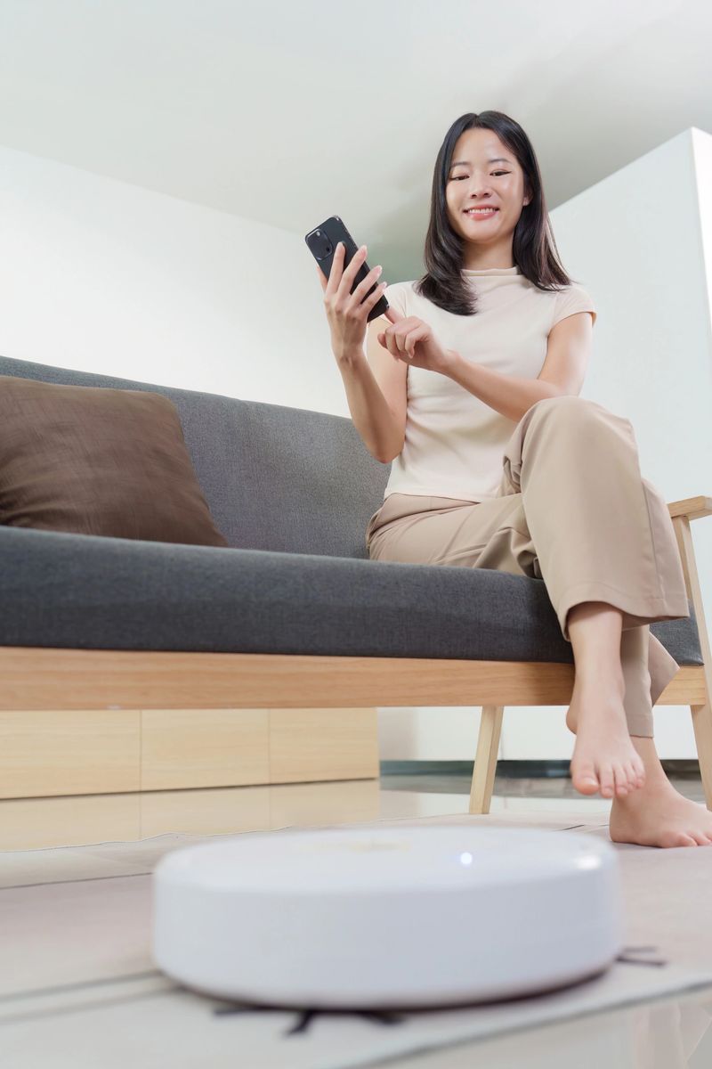 A young woman uses her smartphone to operate a robotic vacuum cleaner in her stylish living room. Emphasizing smart home convenience.
