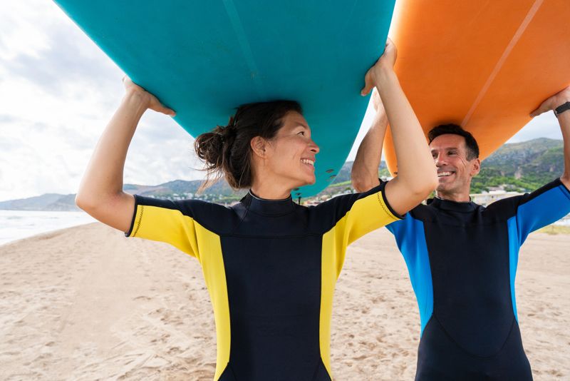 Mature man and asian woman in wetsuits stand on sunny sand, smiling and carrying large surfboards overhead as they prepare for a surf lesson in ocean waves