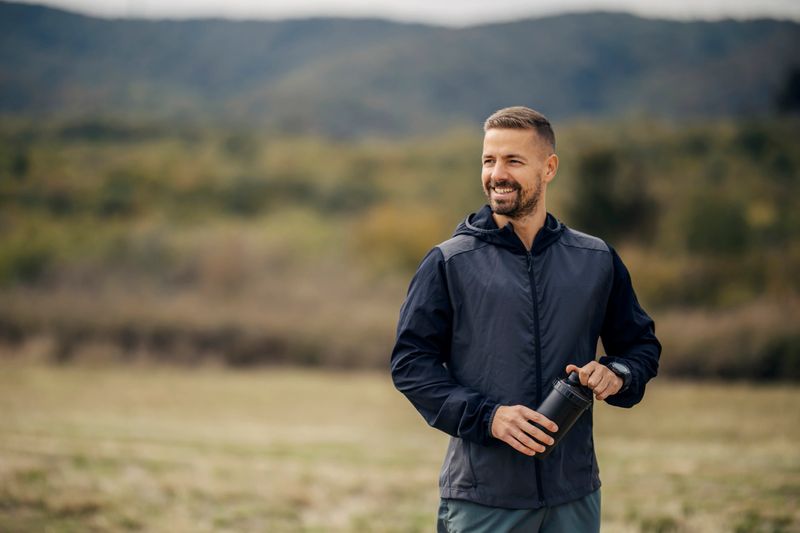 Smiling man enjoying nature, holding a water bottle after a workout or run in the park, staying hydrated