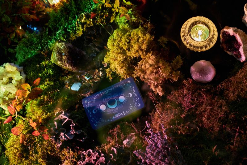 Colorful tarot cards lie on lush moss, surrounded by various crystals and candles in a tranquil forest. scene glows softly as dusk falls, creating a magical atmosphere.