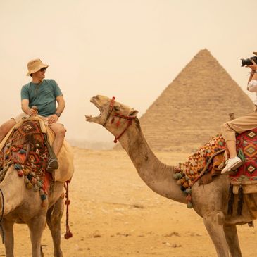 Egypt cairo Giza Pyramids tour packages holiday package, Two tourists on camels near a pyramid, one taking a photo, the other looking amused.