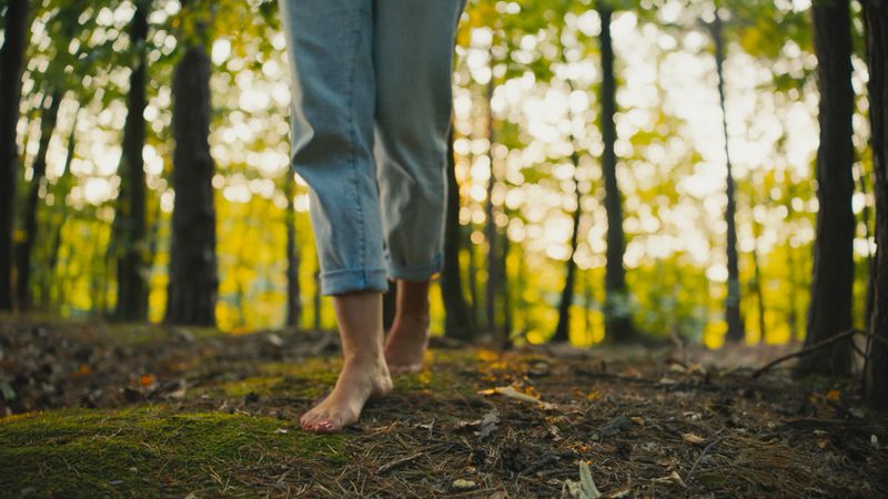 Mid-adult Caucasian woman walking barefoot on a forest floor, enjoying the tranquil atmosphere with sunlight filtering through the trees. The serene setting provides a peaceful escape amidst nature.