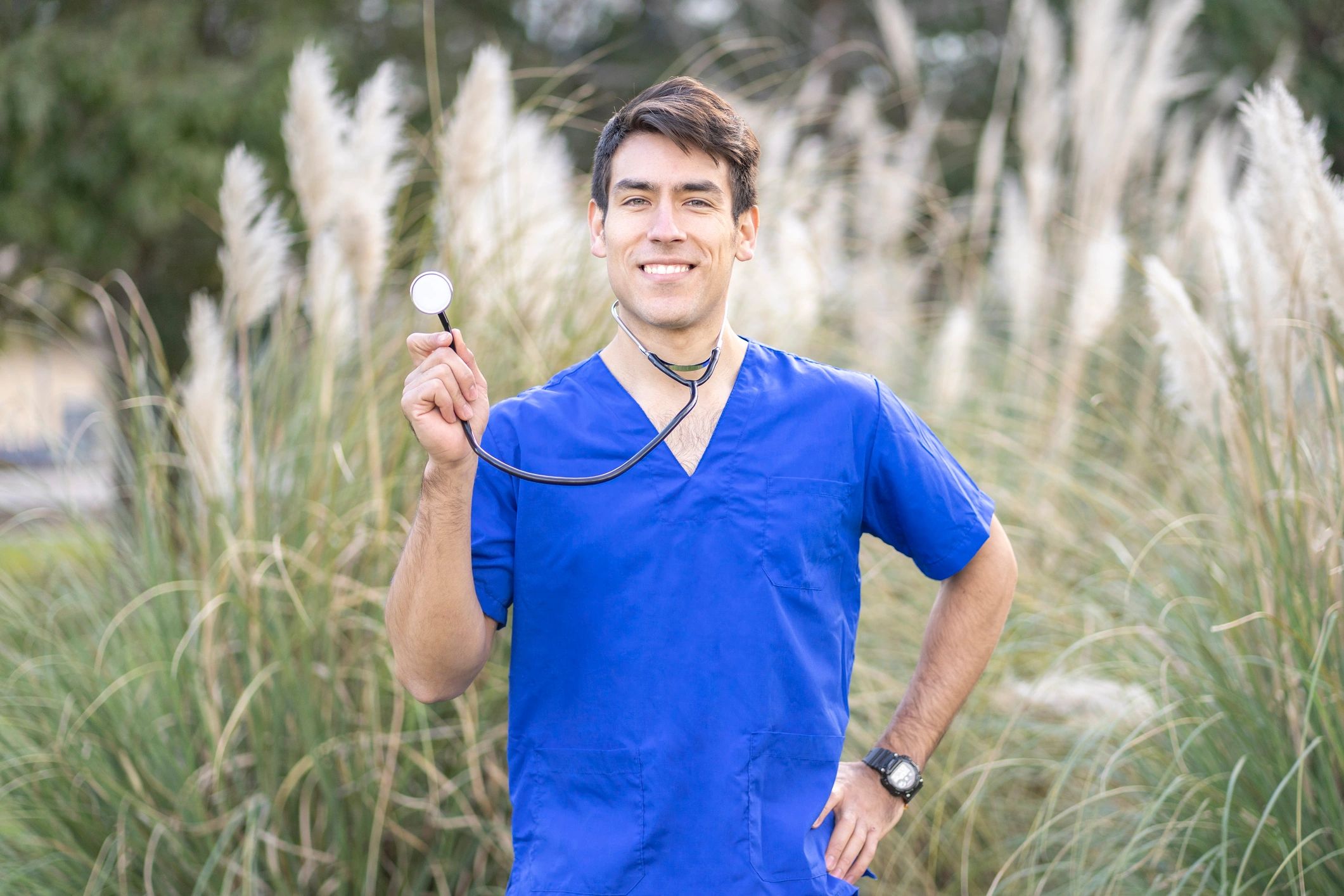 Smiling healthcare professional in blue scrubs holding a stethoscope outdoors.
