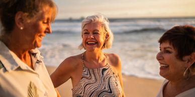 Three older adults happily chatting on a beach at sunset.